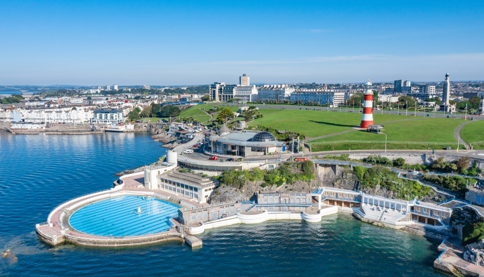 Aerial view of Plymouth Hoe showing Tinside Lido, the coastline and Smeaton’s Tower lighthouse under a clear blue sky.